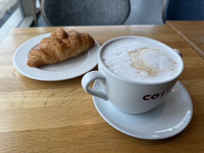 A cappuccino in a Costa Coffee cup served on a wooden table alongside a golden croissant on a white plate.