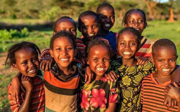 Group of nine smiling African children posing closely together outdoors in a grassy field.