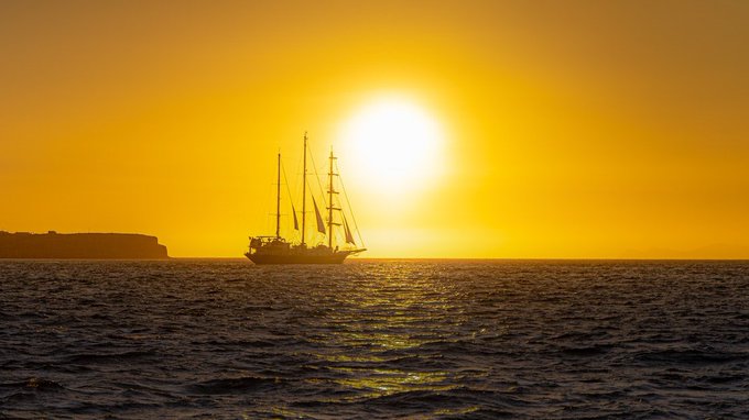 A silhouette of a tall ship with multiple masts sailing on the ocean at sunset, with the sun low in a vibrant orange sky and a faint coastline in the distance.