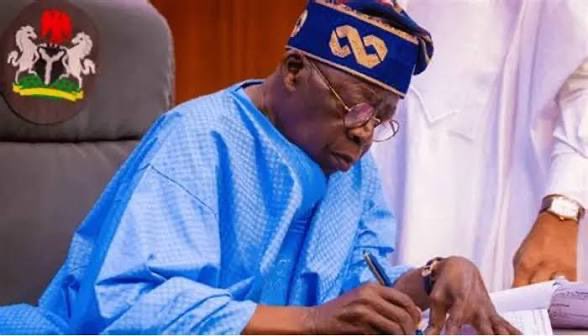 The image shows President Bola Ahmed Tinubu, identifiable by his signature blue traditional attire and cap, signing a document while seated at a desk, with the Nigerian coat of arms visible in the upper left corner—visually representing the moment he signs an executive order regulating cryptocurrencies and virtual assets.