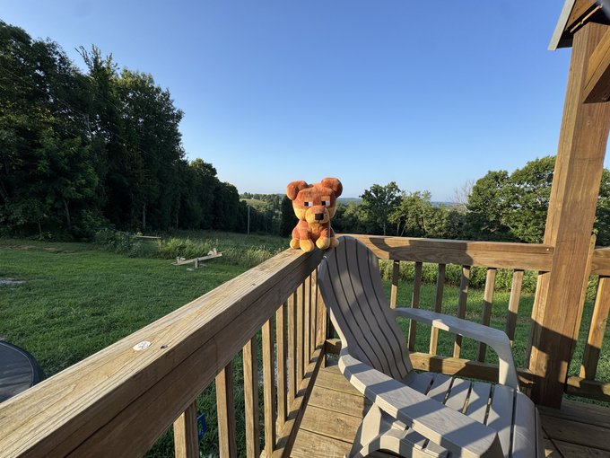 A small brown teddy bear figurine perched on the wooden railing of a deck, with a gray Adirondack chair and a scenic green landscape in the background.