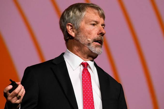 Michael Saylor, wearing a dark suit and red polka-dot tie, speaks on stage while holding a clicker, shown in connection with news that his company Strategy sold 1,638 BTC for $104 million.
