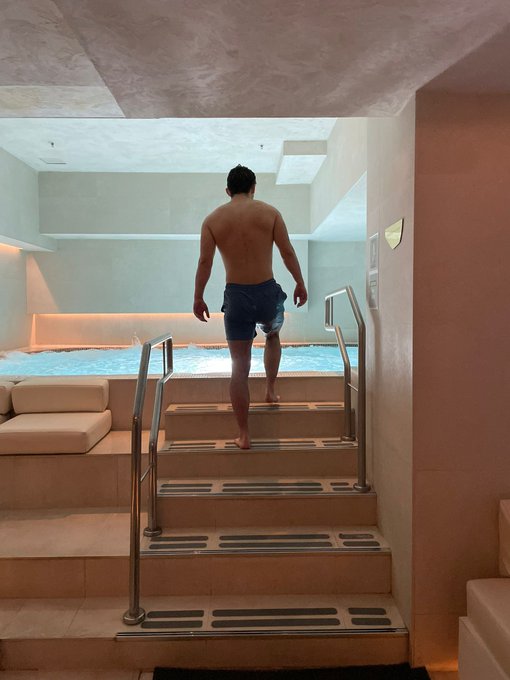 A photo of a shirtless man wearing blue swim trunks walking up steps away from a glowing indoor pool in a modern spa setting.