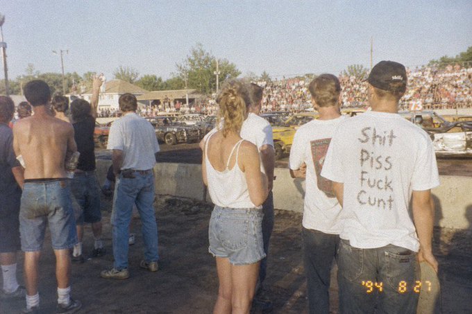 A vintage photograph from '94 8 27' shows a crowd at what appears to be a demolition derby, with a man in the foreground wearing a white t-shirt with handwritten words 'Shit Piss Fuck Cunt'.