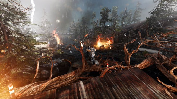A first-person view from a video game showing a chaotic forest battlefield with explosions, fallen trees, and a soldier in the distance.