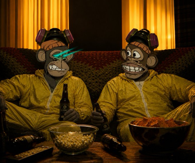 Two cartoon monkey characters in yellow hazmat suits and gas masks sitting on a couch, holding Guinness beer bottles, with bowls of popcorn and chips on the table in front of them.