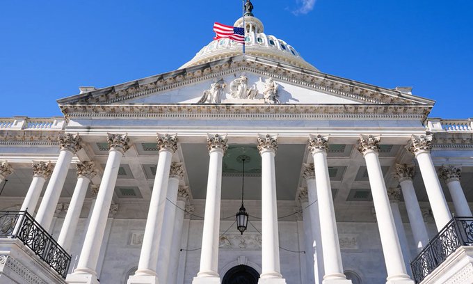 The image shows the U.S. Capitol Building's facade with its iconic white columns, pediment sculpture, and American flag flying atop the dome against a clear blue sky—representing the U.S. Senate chamber where the Crypto Clarity Act was absent from today's legislative agenda.