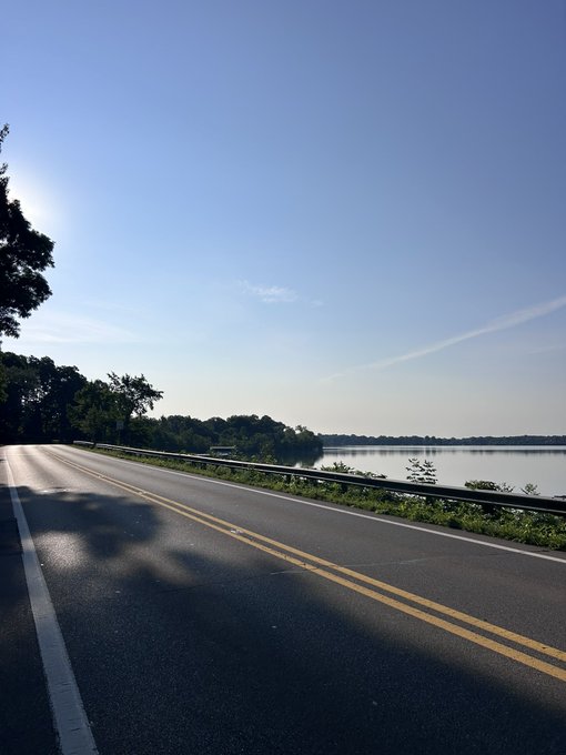 An empty two-lane road runs alongside a calm lake under a clear blue sky, with green trees lining the left side of the sunlit road.