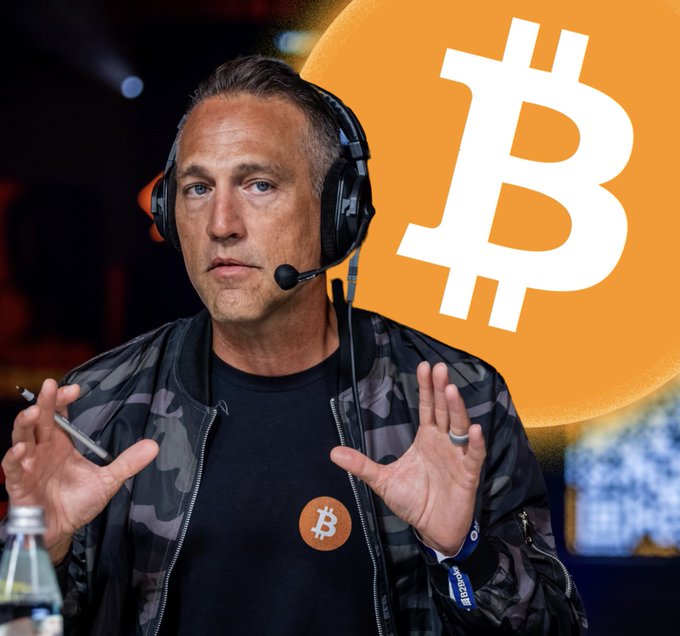 The image shows a man wearing sunglasses standing near a computer screen with a Bitcoin logo displayed at the top right corner of the screen. In the background, there's a sign that says "MARK MOSS SAID 1 BITCOIN WILL BE WORTH $400,000,000 BY 2050". The text on the sign indicates that the Bitcoin value will be doubled in the next six months if it reaches its current value of $4,000,000.