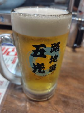 A frothy mug of beer with Japanese characters reading "五光 鶏地酒" (Gokou Chicken Local Sake) on the glass, sitting on a wooden table.