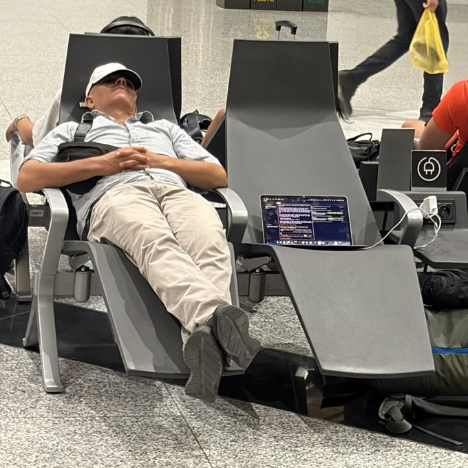 A man in a white cap sleeps soundly across airport seating while his laptop screen displays an AI-generated image of a colorful building, humorously illustrating the "fire off a prompt and go to sleep" workflow of letting generative AI do the work.