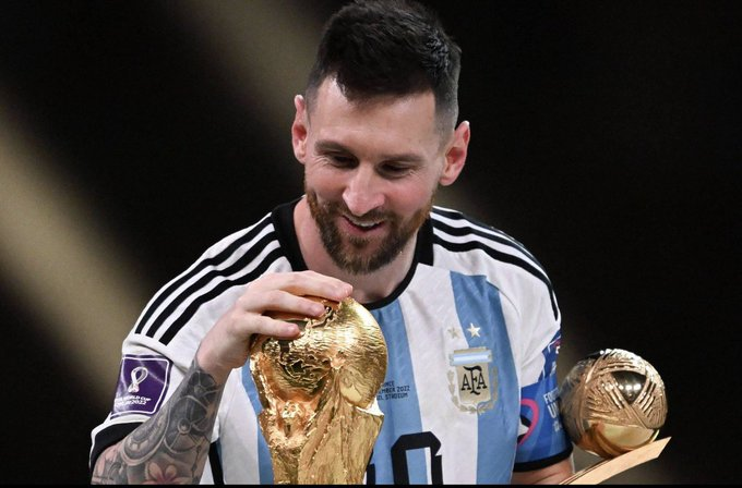 Soccer player Lionel Messi, wearing the Argentina national team jersey, smiles as he kisses the FIFA World Cup trophy while holding a Golden Ball award.