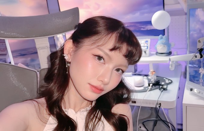 A close-up selfie of a young woman with dark hair, bangs, and dangling earrings, posing in a brightly lit room with shelves displaying figurines in the background.
