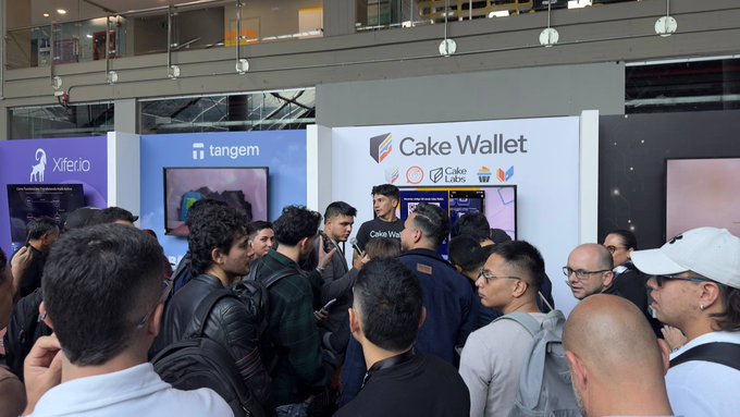 A crowded booth at what appears to be a tech or crypto conference, with people gathered around a display featuring visible branding for 'Xferio', 'tangem', and 'Cake Wallet'.