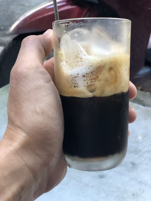 A hand holds up a tall glass of dark Vietnamese iced coffee (cà phê đá) with a metal straw, the rich brown liquid topped with a frothy layer of cream and ice. The casual, on-the-go presentation of the coffee fits the post's "morning ritual" vibe of enjoying a quick cà phê đá while checking in for updates on the QUIP network.
