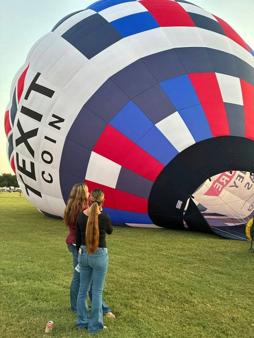 A TEXITcoin-branded hot air balloon dominates a grassy field at the Plano Balloon Festival, where two women stand gazing up at its massive red, white, and blue patterned envelope as it prepares for launch. The bold "TEXIT COIN" lettering on the balloon's side mirrors the post's theme, visually bringing the cryptocurrency brand to life as it "takes flight" in the community.