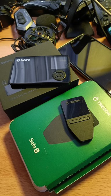 A close-up shot of two hardware cryptocurrency wallets: a black SafePal device resting on its box and a Trezor Model T sitting on top of a green Safe 3 box, with various cables visible in the background.