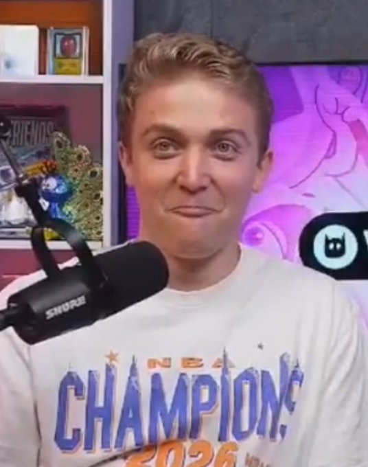 A young man with short blonde hair sits in front of a microphone, wearing a white t-shirt that reads 'NBA CHAMPIONS 2024' with stars, while making a pursed-lip expression.