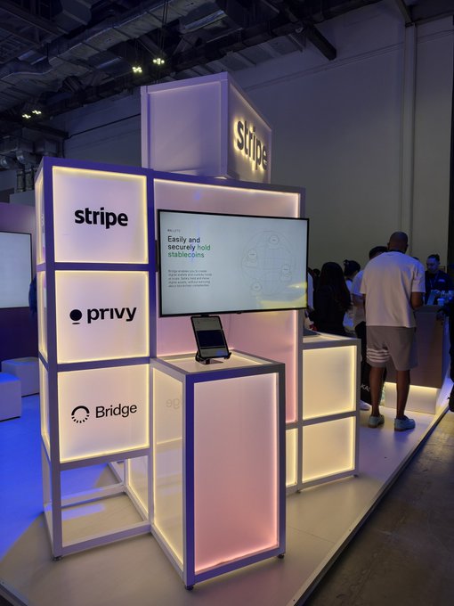 A brightly lit booth at an exhibition displaying logos for Stripe, Privy, and Bridge on stacked illuminated cubes, with a screen showing 'Privy Easily and securely hold stablecoins'.
