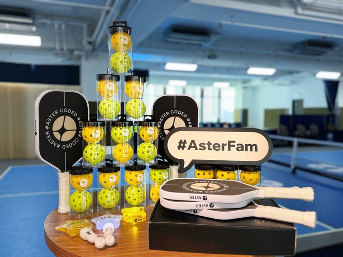 A display of pickleball equipment is arranged on a table, featuring stacked tubes of yellow and green balls, two paddles, and signs that read '#ASTE... MASTER CODED BY ASTER' and '#AsterFam'.