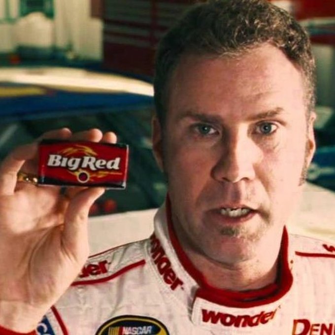 A close-up of NASCAR driver Ricky Bobby, played by Will Ferrell, holding up a tin of 'Big Red' chewing tobacco while wearing his racing suit with 'Wonder' and 'NASCAR' logos visible.
