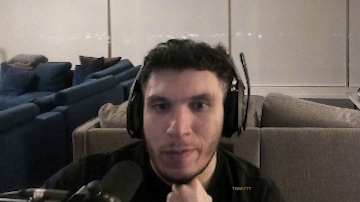 The image features a man wearing headphones with his eyes closed while sitting down. He appears to be smiling or making eye contact as he looks at the camera, possibly posing for a photo or video. The background of the image shows a couch and several chairs arranged in various positions around the room.