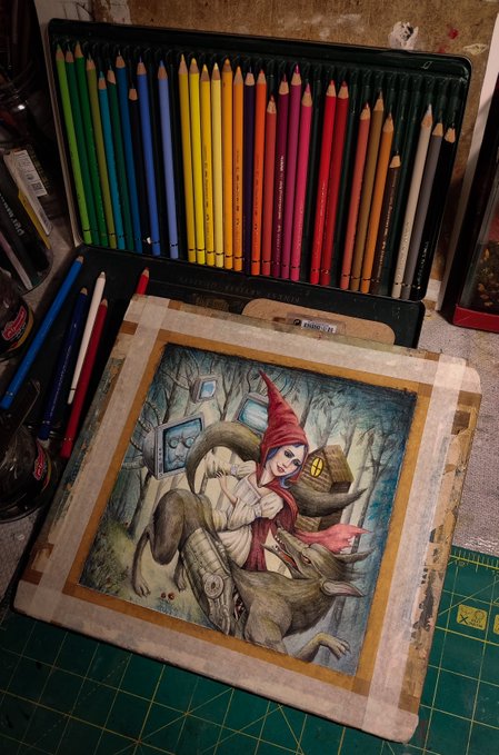 An overhead view of an open tin box of colorful Prismacolor pencils above a detailed colored pencil illustration of a hooded character in a fantasy forest scene, resting on a green cutting mat.