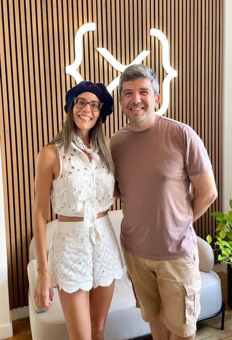 A woman in a white crochet outfit and navy beret poses beside a smiling man in a mauve t-shirt in front of a wooden slat wall featuring an illuminated "M" logo. The image captures a meetup in Paris between the two, tying into the post about an upcoming podcast conversation with Rachel Wolfson.