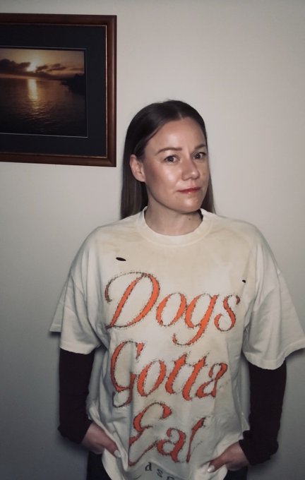 A young woman with brown hair wears a distressed, oversized cream t-shirt with the faded red text 'Dogs Gotta Eat', layered over a long-sleeve black shirt.