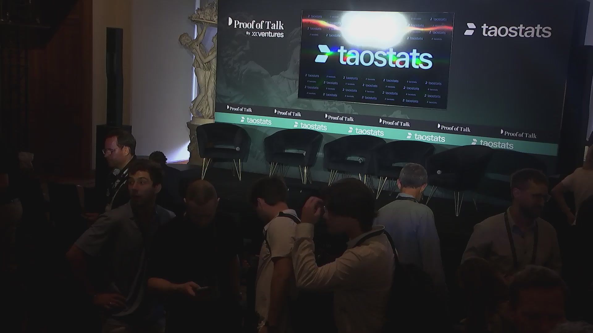 The image shows a group of people gathered around a large screen displaying information about TOSATs, which stands for "Tosat Statistics". The crowd appears engaged with the content being presented by the speaker or presenter at this event. There are several chairs positioned in front of the screen and some individuals can be seen standing nearby. A person is holding a cell phone, possibly taking notes or capturing photos related to the presentation.