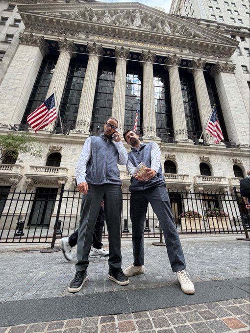 Two men in matching gray outfits posing in front of the New York Stock Exchange building with American flags displayed.