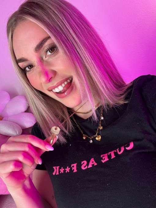 A young woman with long hair featuring purple highlights smiles at the camera while wearing a black t-shirt with 'CUTE AS F**K' printed on it, bathed in pink lighting.