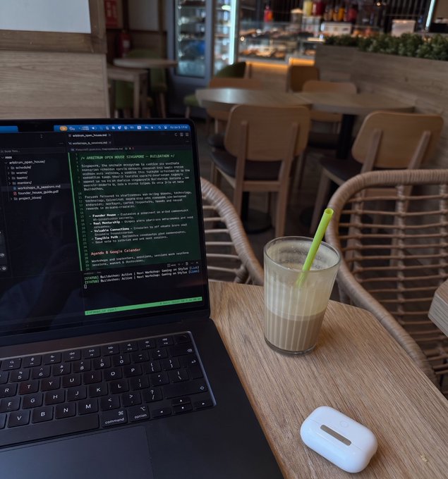 A laptop displays code for an "Arbitrum Open House Singapore — Buildathon" project alongside an iced coffee at a café, with AirPods resting nearby—capturing the essence of a developer's casual work session to build and refine their summer side project.