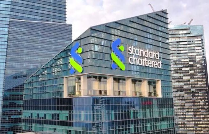 A modern glass office building featuring the Standard Chartered bank logo with its distinctive green and blue interlocking symbol on the facade.