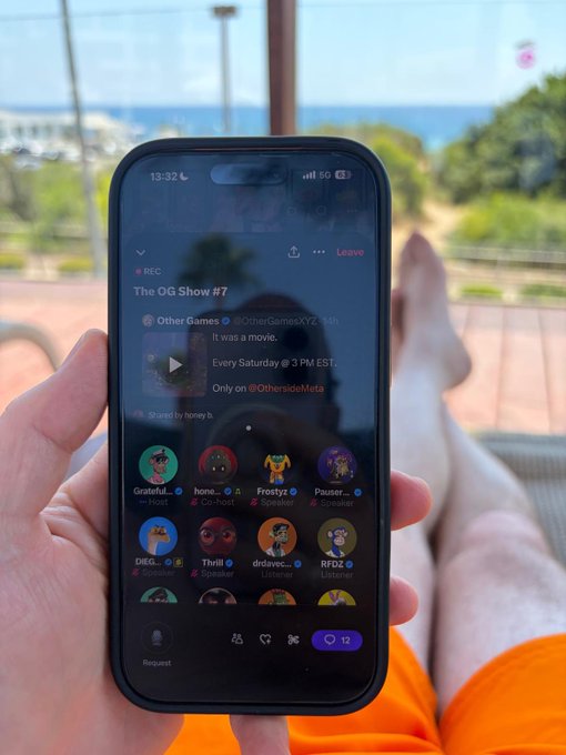A person's hand holding a smartphone displaying a Twitter Space called "The OG Show #7" hosted by "Other Games" (@OtherGamesXYZ), with 12 participants, set against a background of an ocean view.