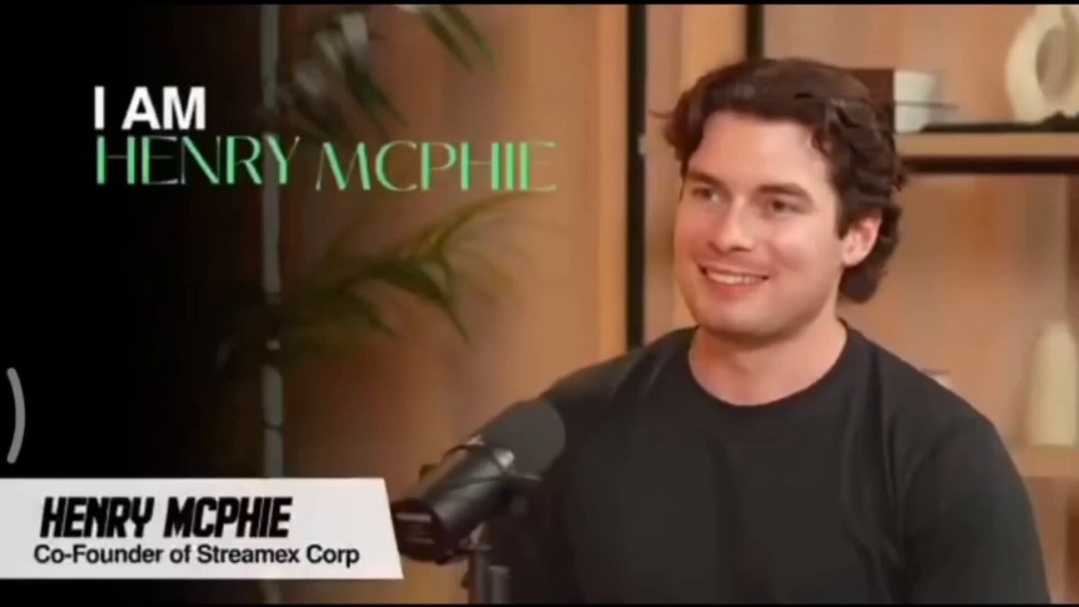 The image features a man wearing a black shirt who appears to be smiling at the camera while speaking into a microphone, possibly giving an interview or hosting a podcast. The background includes a bookshelf filled with various items such as books and vases. There is also text in the image that reads "I am Henry Mchrie Co-Founder of StreamX Corp".