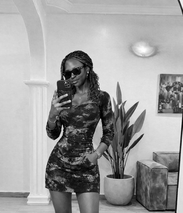 A black and white mirror selfie of a woman wearing sunglasses and a patterned mini dress, standing next to a potted plant.