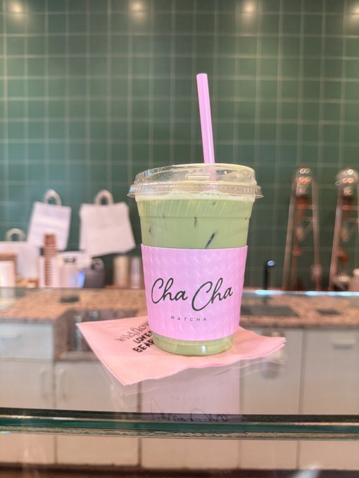 A green iced matcha drink in a clear cup with a pink 'Cha Cha MATCHA' sleeve and pink straw sits on a pink napkin atop a glass counter in a tiled cafe.