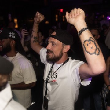 A bearded man in a white t-shirt and backwards black cap raises his arms in celebration. He has a recognizable flower-shaped tattoo on his forearm and wears a lanyard, appearing to be in a crowd at a concert or event.