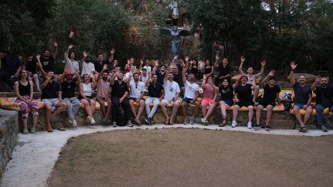 A group photo of around twenty people gathered outdoors at sunset in front of a large statue with outstretched arms, with several participants raising their hands in celebration.