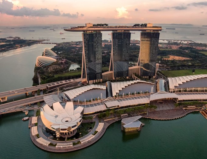 An aerial view of Marina Bay Sands in Singapore at sunset, showing the three-tower hotel, the lotus-shaped ArtScience Museum, and waterfront surroundings.
