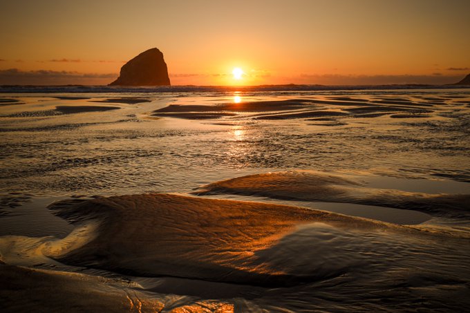 A sunset scene at the beach shows golden light reflecting off rippled wet sand, with a large rock formation silhouetted on the horizon.
