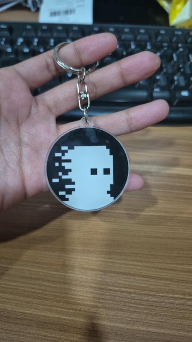 A hand holds a circular acrylic keychain featuring a black and white pixel-art skull design above a keyboard.