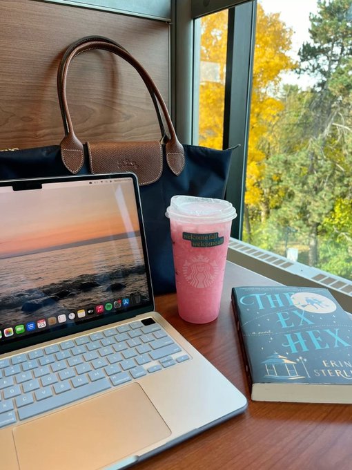 A laptop displaying a sunset wallpaper sits on a wooden table next to a Starbucks pink drink in a reusable sleeve with text reading 'Welcome back autumn!', alongside a copy of the book 'The Ex Hex' by Erin Sterling, with autumn trees visible outside the window.