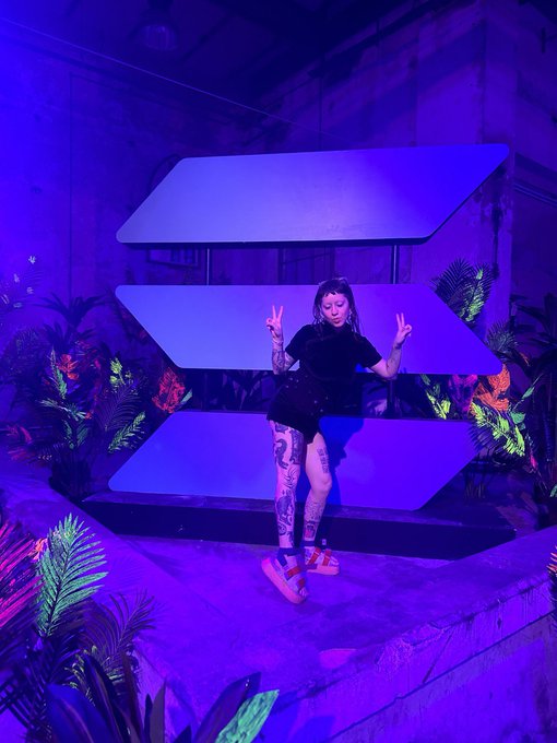 A woman with tattoos wearing a black top and shorts stands in front of a large blue geometric Solana (SOL) logo, posing with peace signs under purple lighting.