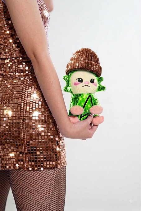 A person in a copper-sequined dress and fishnet stockings holds a small green plush toy wearing a brown beanie, referencing the "Chog" meme associated with RobinhoodCrypto branding.