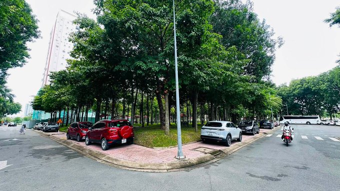 A street-level panoramic view of an intersection or parking area lined with lush green trees, featuring a few parked cars (red and white SUVs) and a bus in the distance.