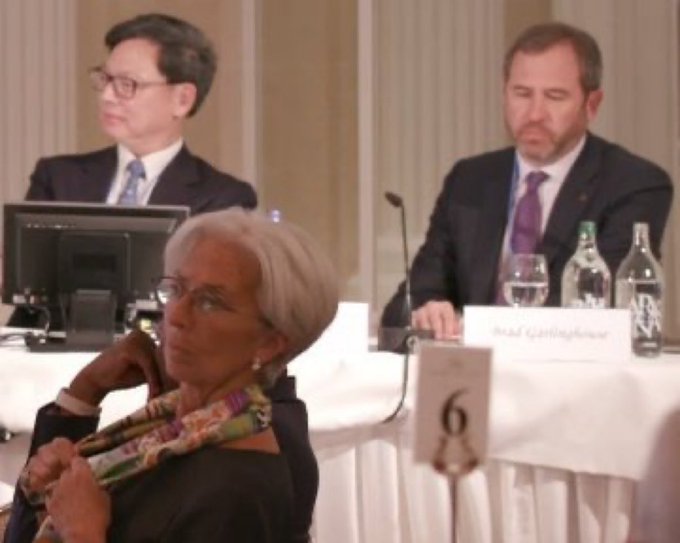 A formal conference setting shows Christine Lagarde, ECB President, seated at a table in the foreground with a colorful scarf, while behind her two men in suits appear at a panel desk, one looking at a monitor and another reviewing documents near a nameplate reading "Wulf Guttering" and a table number "6." The image relates to the post "$XRP ECB" by depicting a meeting of European Central Bank officials, a context where XRP and cryptocurrency regulatory discussions may occur.