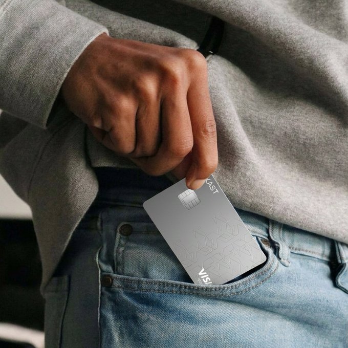 A close-up of a person in a gray sweatshirt and blue jeans holding a silver Visa credit card and sliding it into their front pocket.
