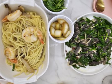 An overhead view of a meal featuring pasta with shrimp in a white baking dish, accompanied by a plate of sautéed beef with green onions, side dishes of greens and pickled radish, and chopsticks resting on the counter.